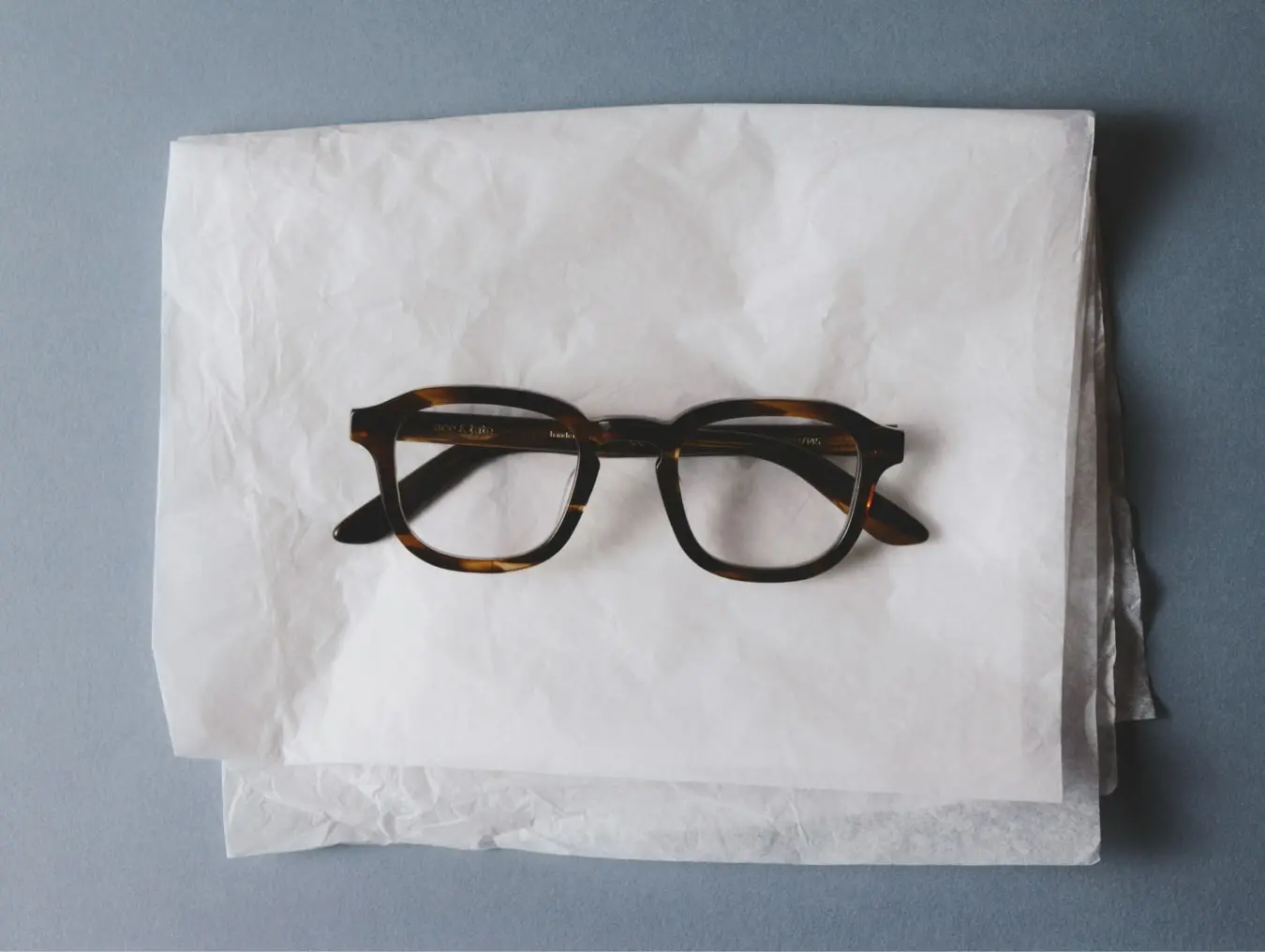 Ace & Tate | A pair of thin, round metal-framed glasses nesting in crumpled craft paper inside a small cardboard box.